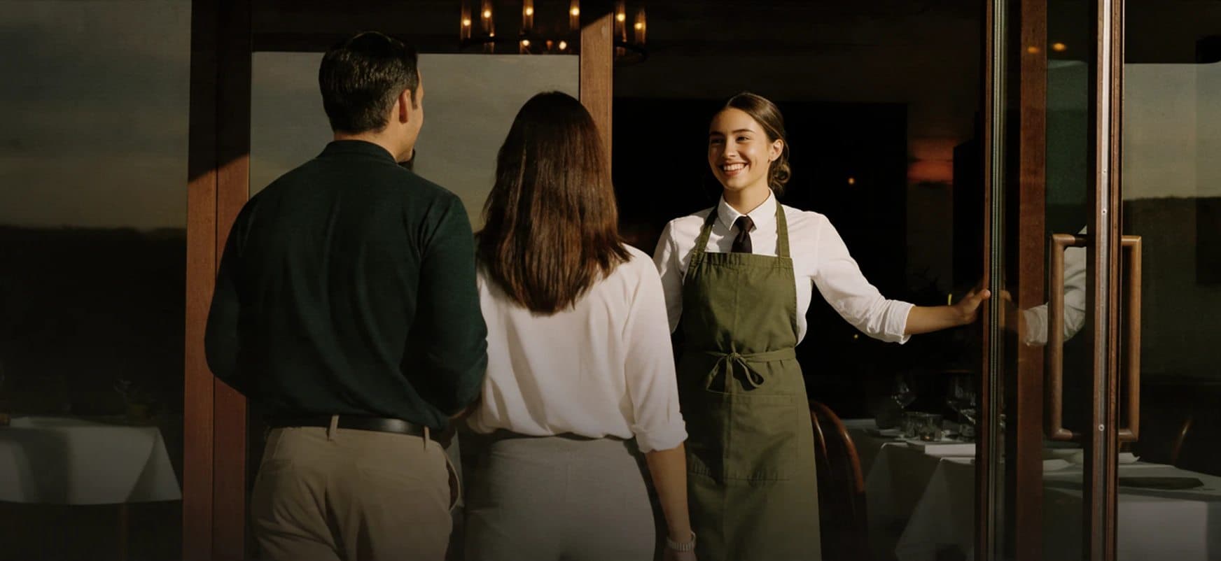 Una anfitriona sonríe y sostiene la puerta de un restaurante mientras recibe a una pareja que llega.