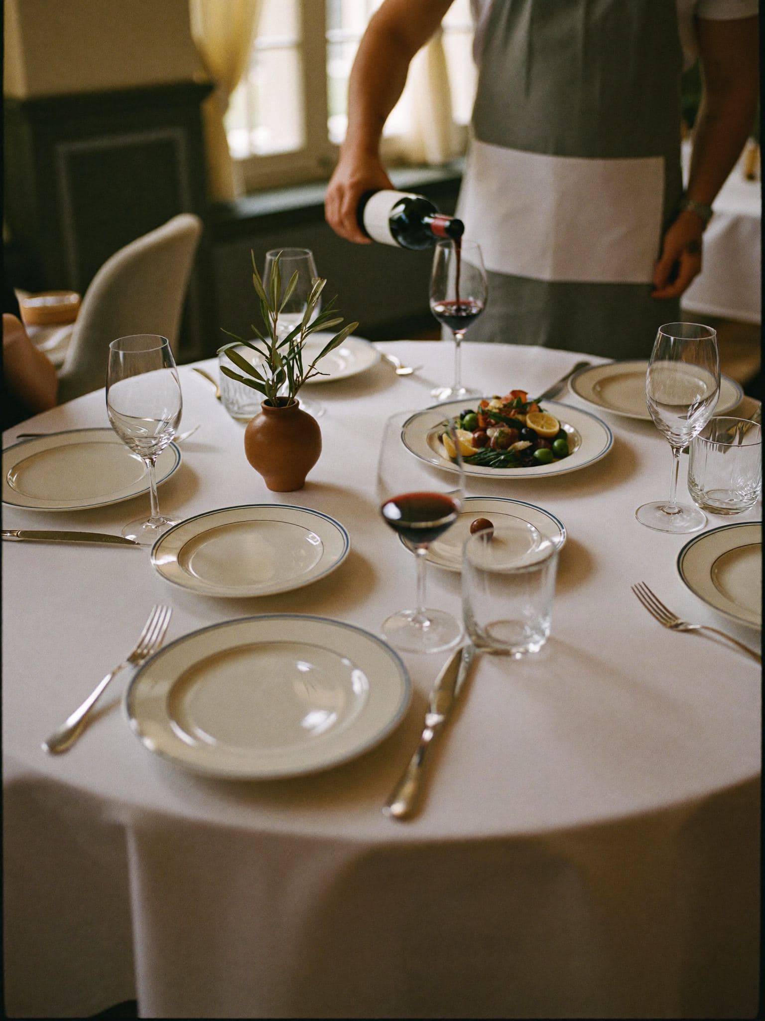 Una mesa de un restaurante fino con mantel blanco, copas de cristal y un camarero sirviendo vino tinto.